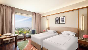 A hotel room with two beds, a dining table, a chair, a small table with a vase, and a view of the city.