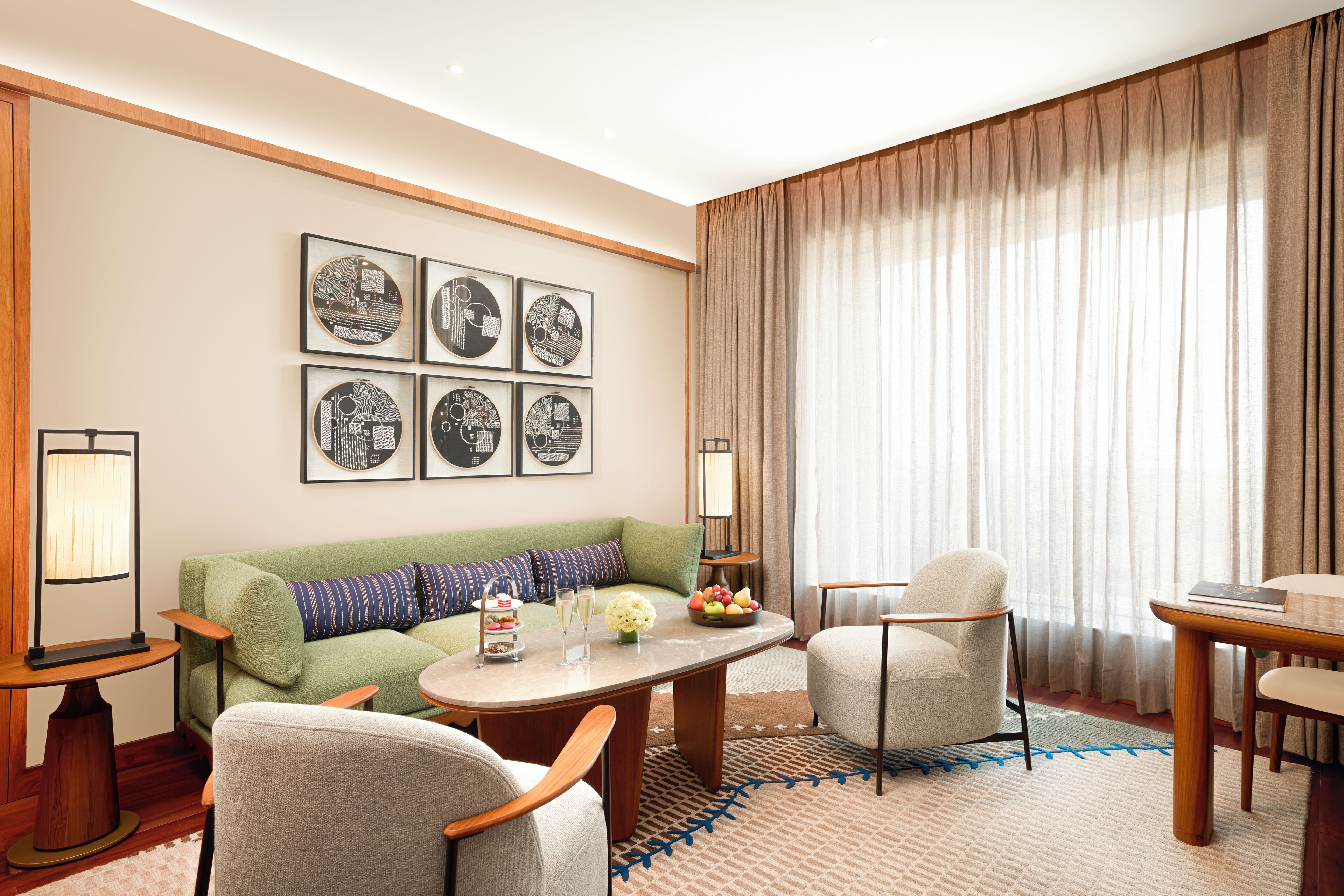 A hotel room with a green sofa, two armchairs, a round coffee table, and a dining table. There are framed pictures on the wall and a window with curtains.
