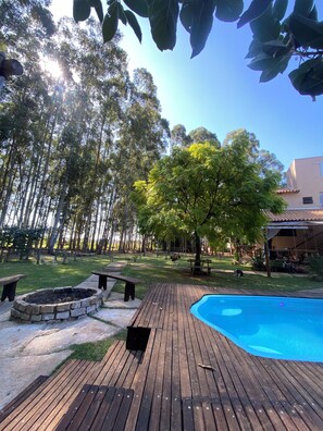 A wooden deck with a swimming pool, surrounded by a grassy area with trees and a stone fire pit. - Hotel e Pousada do Bosque (Boa Esperanca)