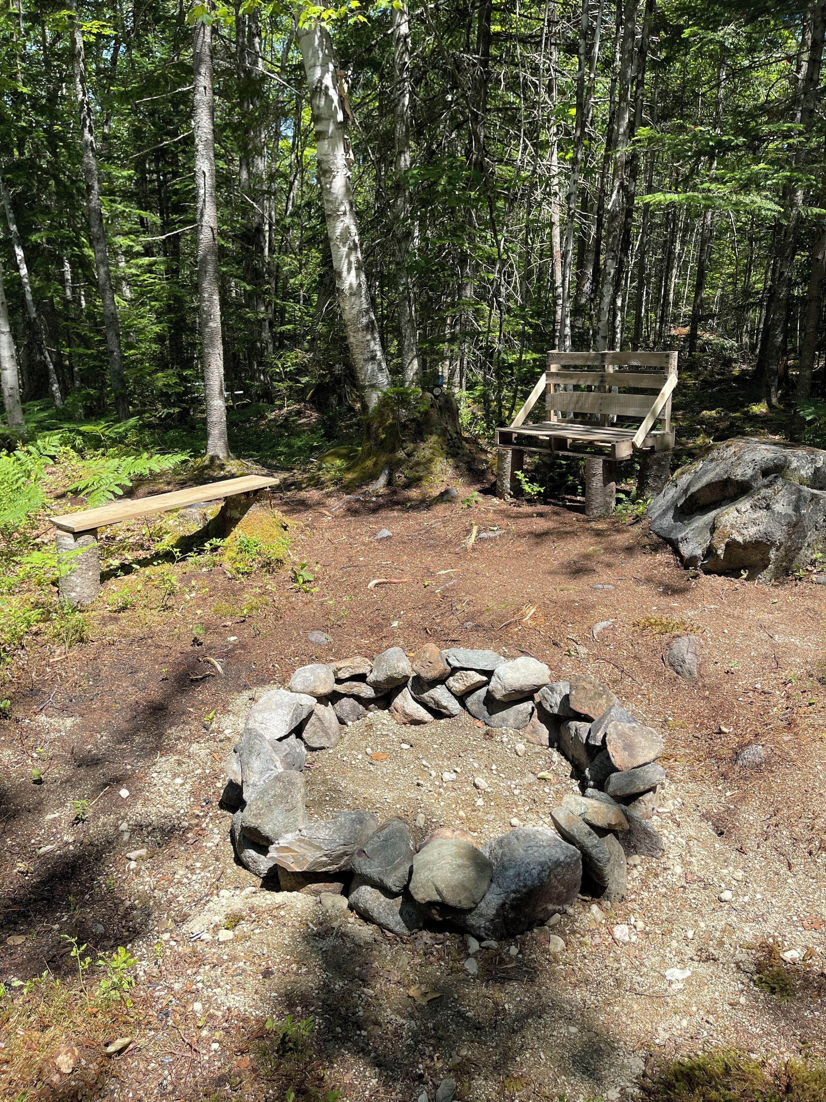 Een camping met een vuurkorf van steen, een houten bank en een picknicktafel, omgeven door bomen.