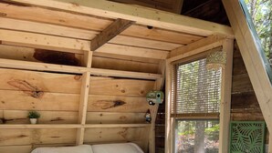 Ein Schlafzimmer einer Holzhütte mit einem Bett, einem Nachttisch, einem kleinen Regal mit einer Pflanze und einem Fenster mit Blick auf Bäume.
