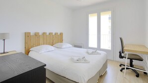 A bedroom with a wooden headboard, a bed with white linens, a desk with a chair, and a window with blinds. - Appartement des Chênes - Welkeys (Anglet)