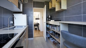 A compact kitchen with a built-in dishwasher, a coffee maker, and open shelves with dishes. - Appartement des Chênes - Welkeys (Anglet)