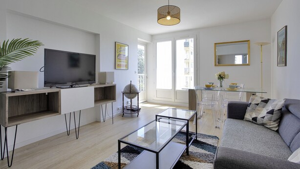 A modern living room with a flat-screen TV, glass coffee table, and a sofa. There is a dining area with a glass table and chairs, and a large window providing natural light. - Appartement des Chênes - Welkeys (Anglet)