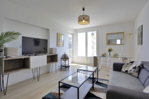 Un salon moderne avec un téléviseur à écran plat, une table basse en verre et un canapé. On y trouve également un coin repas avec une table et des chaises en verre, ainsi qu’une grande fenêtre qui laisse entrer la lumière naturelle.