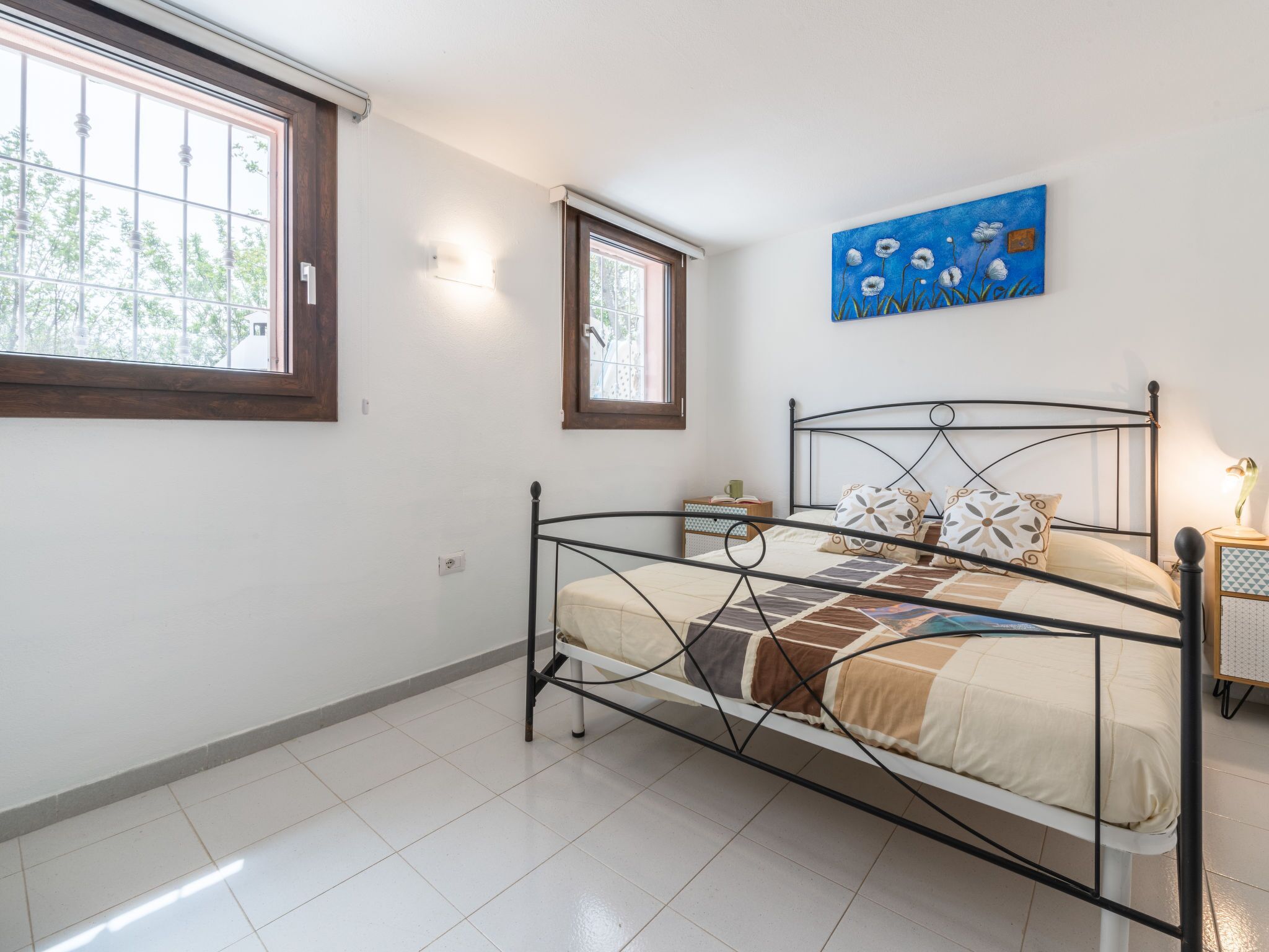 Quarto com estrutura de cama de metal, janela com vista para árvores, quadro na parede e mesa de cabeceira com abajur.