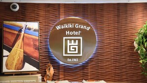 A reception area with a wooden textured wall, a large circular sign reading "Waikiki Grand Hotel Est. 1963", and a framed picture of a paddleboard.