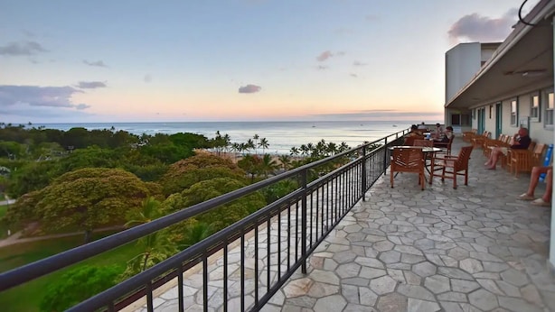 A balcony with stone flooring and metal railings overlooking a beach with palm trees and the ocean.