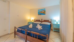 A neatly made bed with blue bedding and two rolled towels, a painting of a coastal scene on the wall, and two bedside tables with lamps.