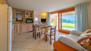 A cozy living room with a sofa and a dining area with a table and chairs. There is a kitchen with cabinets and a refrigerator. A large window offers a view of the outdoors.