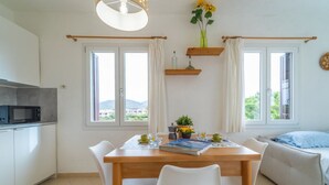 Moderni keittiö, jossa on mikroaaltouuni, ruokapöytä tuoleineen ja sohva. Huoneessa on ikkunat, joissa on verhot, hyllyllä kasvi ja ikkunasta avautuu näkymä ulos.