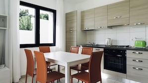 Een moderne keuken met lichte houten kastjes, een witte eettafel met bruine stoelen en een groot raam met witte gordijnen.