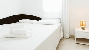 Een net opgemaakt bed met witte lakens en kussens, een nachtkastje met een lamp, en een raam met gordijnen.