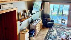 A cozy cabin interior with a wooden ceiling, a welcome sign, a mounted TV, a leather chair, and a large window overlooking a lake.