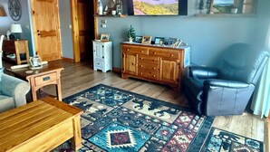 A cozy living room with a wooden ceiling, a large flat-screen TV displaying a scenic view, a leather armchair, a wooden coffee table, and a patterned rug.