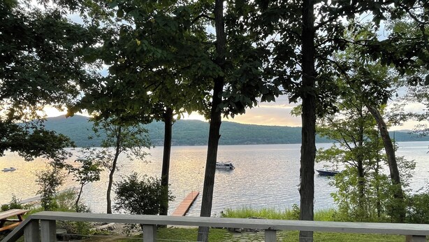 A lakeside view with a wooden deck and railing, surrounded by trees and a distant boat on the water.