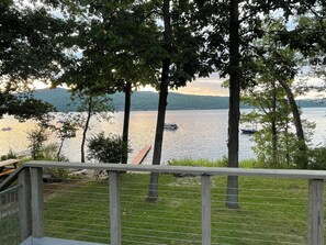 A lakeside view with a wooden deck and railing, surrounded by trees and a distant boat on the water. - Lake front year round home on beautiful Willsboro Bay, Lake Champlain (Willsboro)