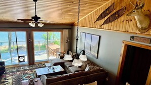 A cozy cabin interior with a wooden ceiling, a large window overlooking a lake, a dining area with a table and chairs, and a mounted deer head on the wall.