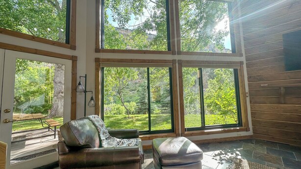 Una habitación amplia con ventanas grandes, un sofá de cuero, un pequeño reposapiés y piso de baldosas de piedra.