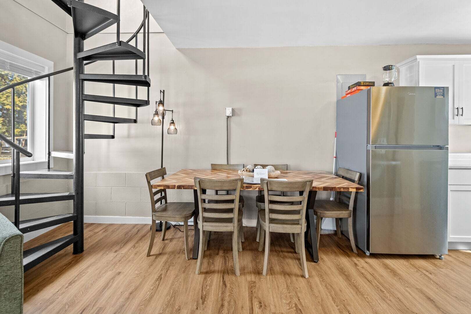 Een moderne keuken met een spiltrap, een eettafel met stoelen, een roestvrijstalen koelkast en een raam met uitzicht op bomen.