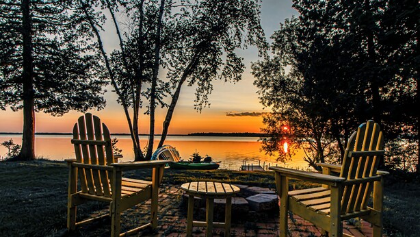 Vista del atardecer con dos sillas Adirondack y una pequeña mesa en un patio de ladrillo junto a un lago.