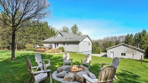 A house with a deck, a fire pit with rocks, and several Adirondack chairs.