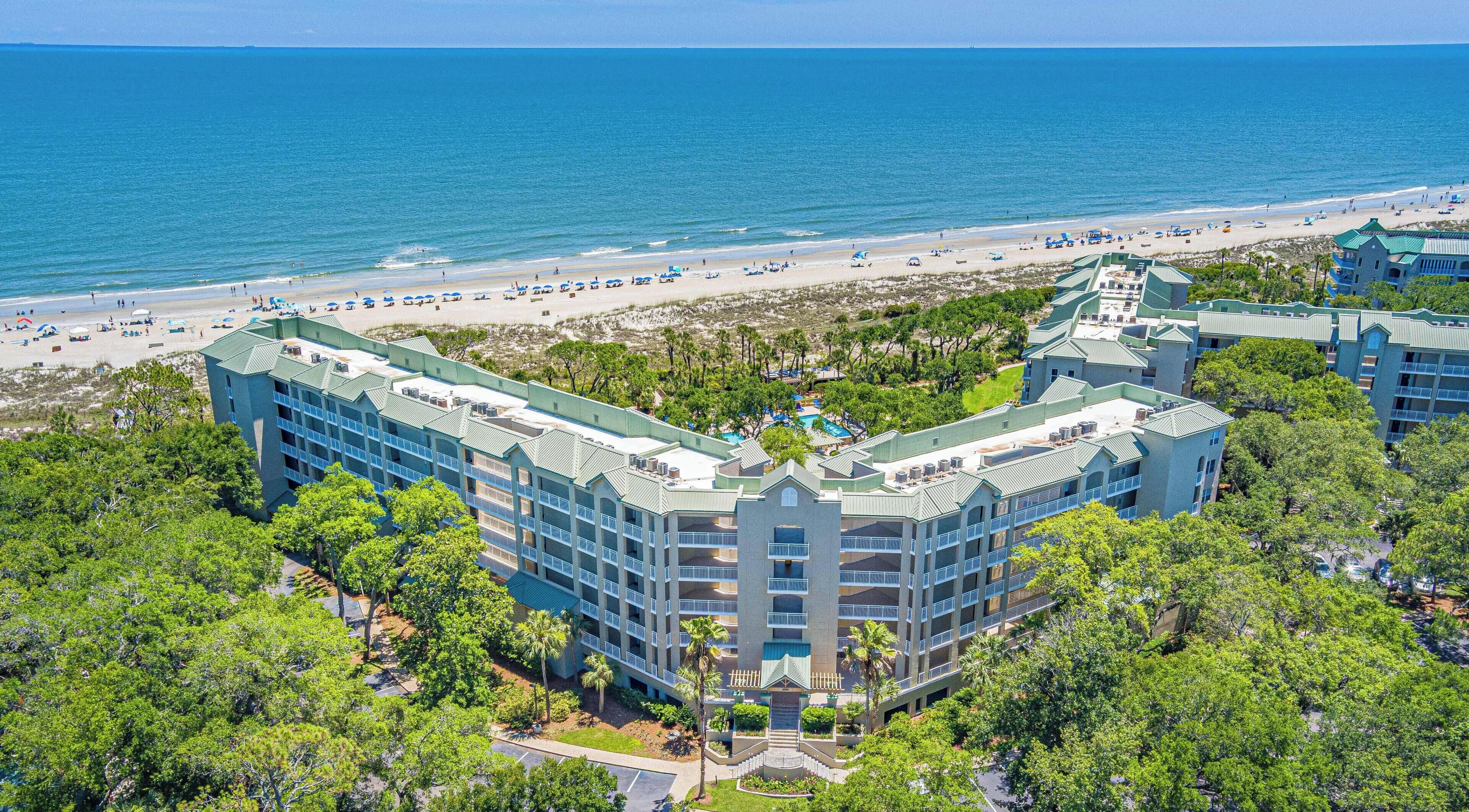 A multi-story hotel with a pool, surrounded by lush greenery and a beach.