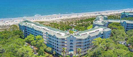 A multi-story hotel with a pool, surrounded by lush greenery and a beach.