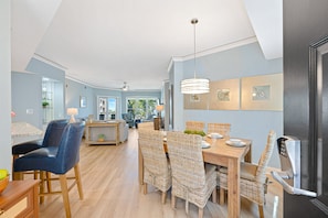 A spacious living area with a dining table, wicker chairs, and a modern kitchen in the background. - Oceanfront Family Villa | Ocean/Pool Balcony Views, Bikes, Complex Pool/Spa (Hilton Head Island)