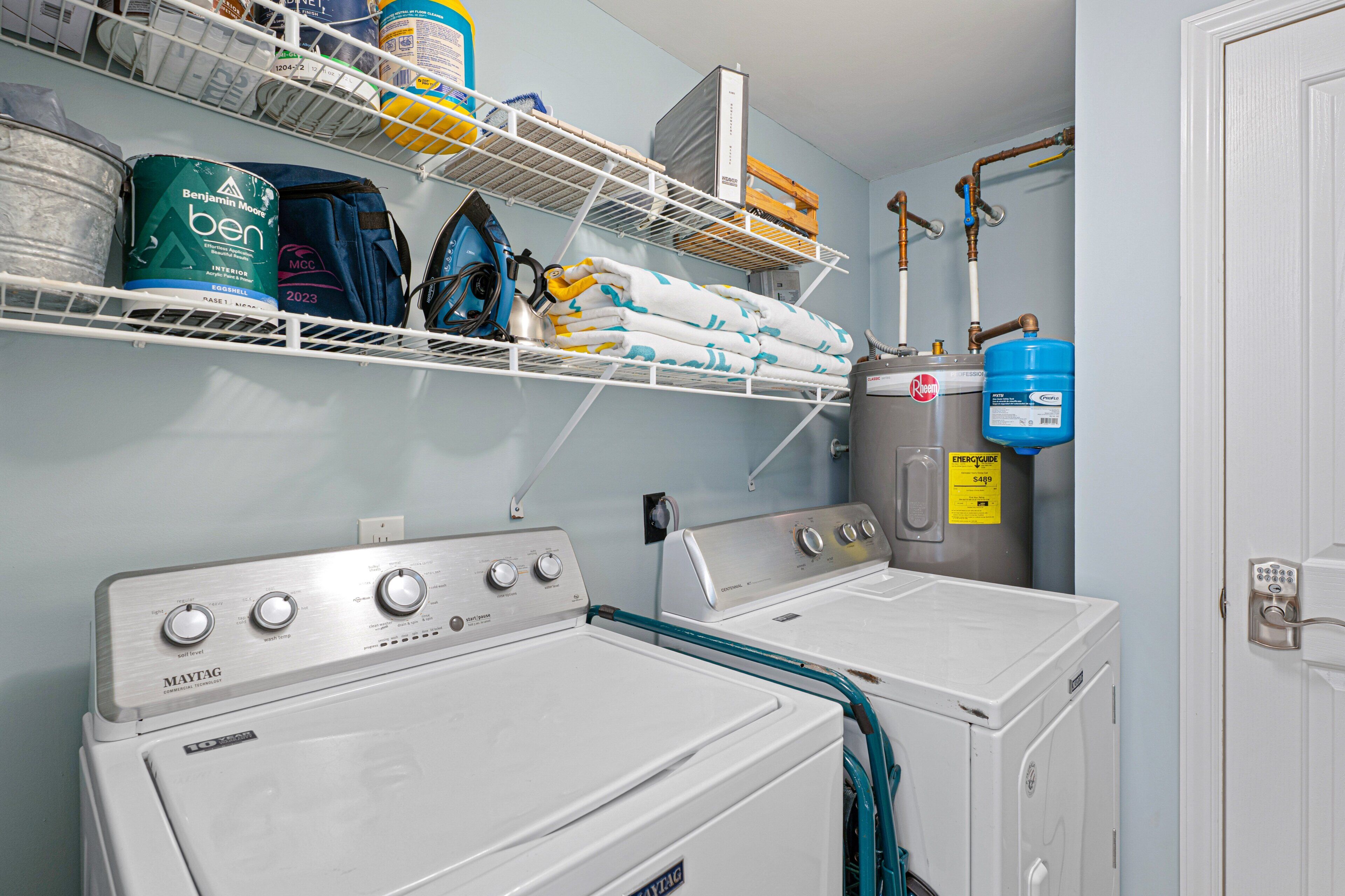 Uma lavanderia com duas máquinas de lavar e secar Maytag brancas, prateleiras com produtos de limpeza e um aquecedor de água.