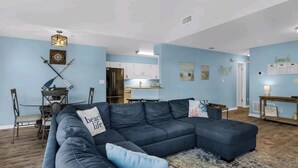 A living room with a sectional sofa, a dining table with chairs, a kitchen area, and a small seating area with a bench. - Seashell Suite! (Destin)