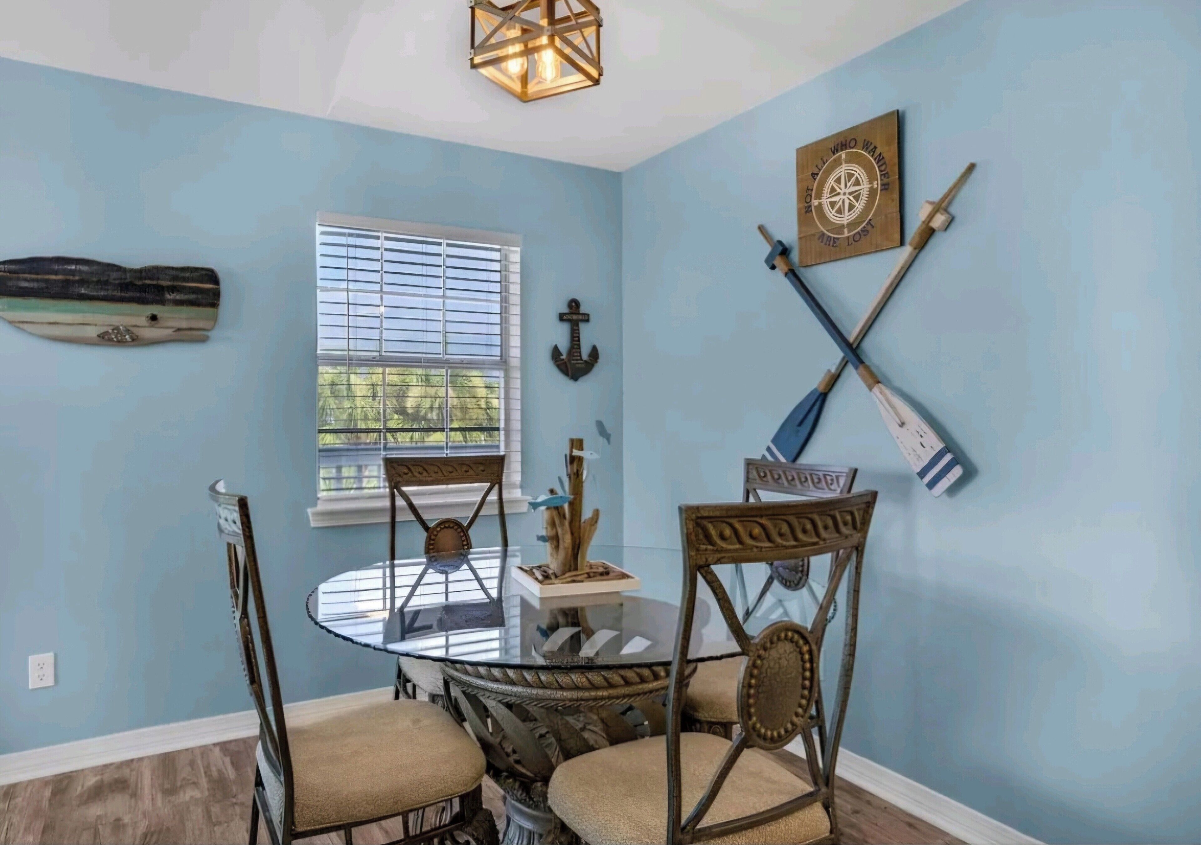 A dining area with a round glass table, four chairs, a wooden ceiling light, and nautical-themed wall decor.