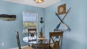A dining area with a round glass table, four chairs, a wooden ceiling light, and nautical-themed wall decor. - Seashell Suite! (Destin)