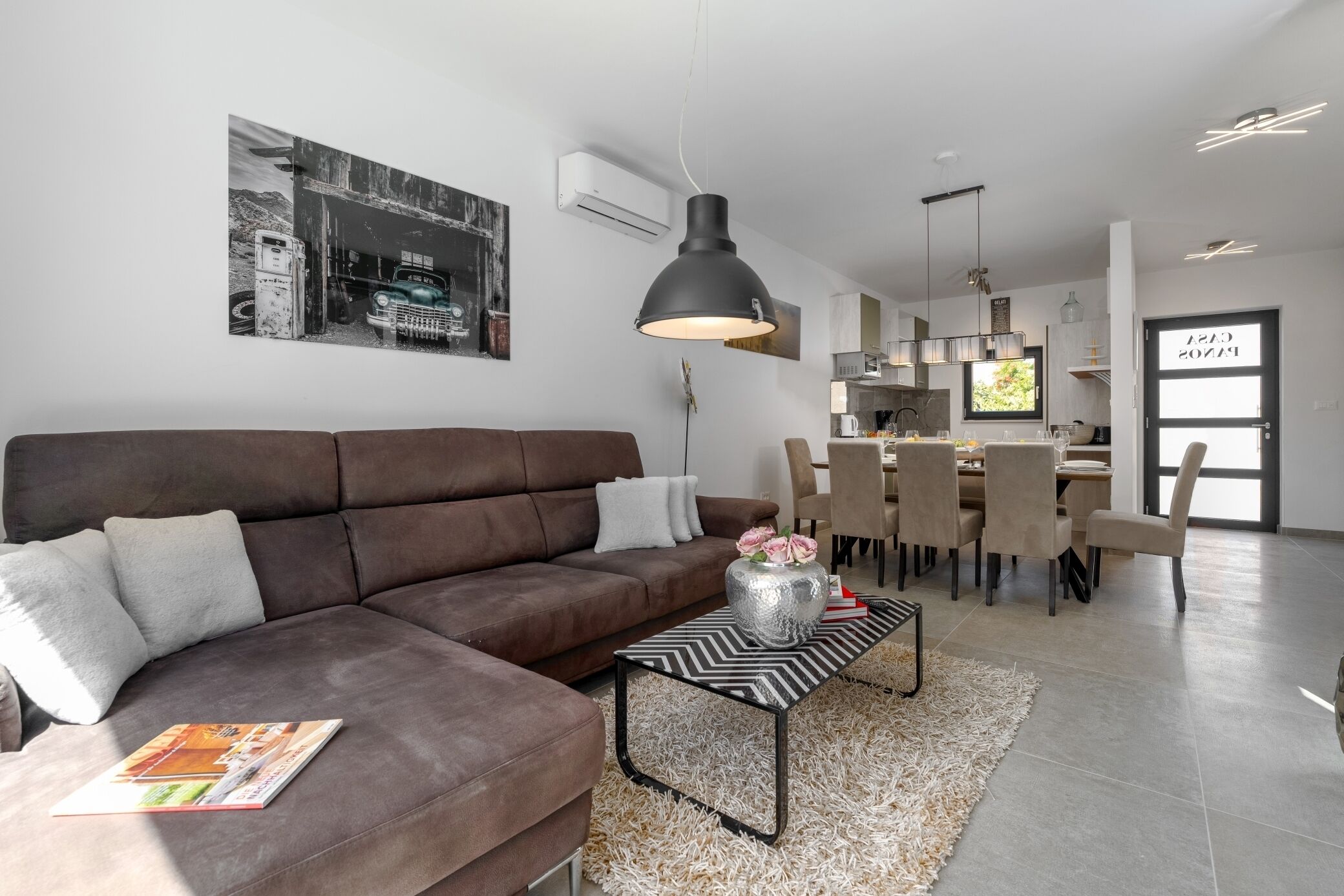 A modern living room with a brown sofa, a black and white coffee table, and a dining area with a chandelier.