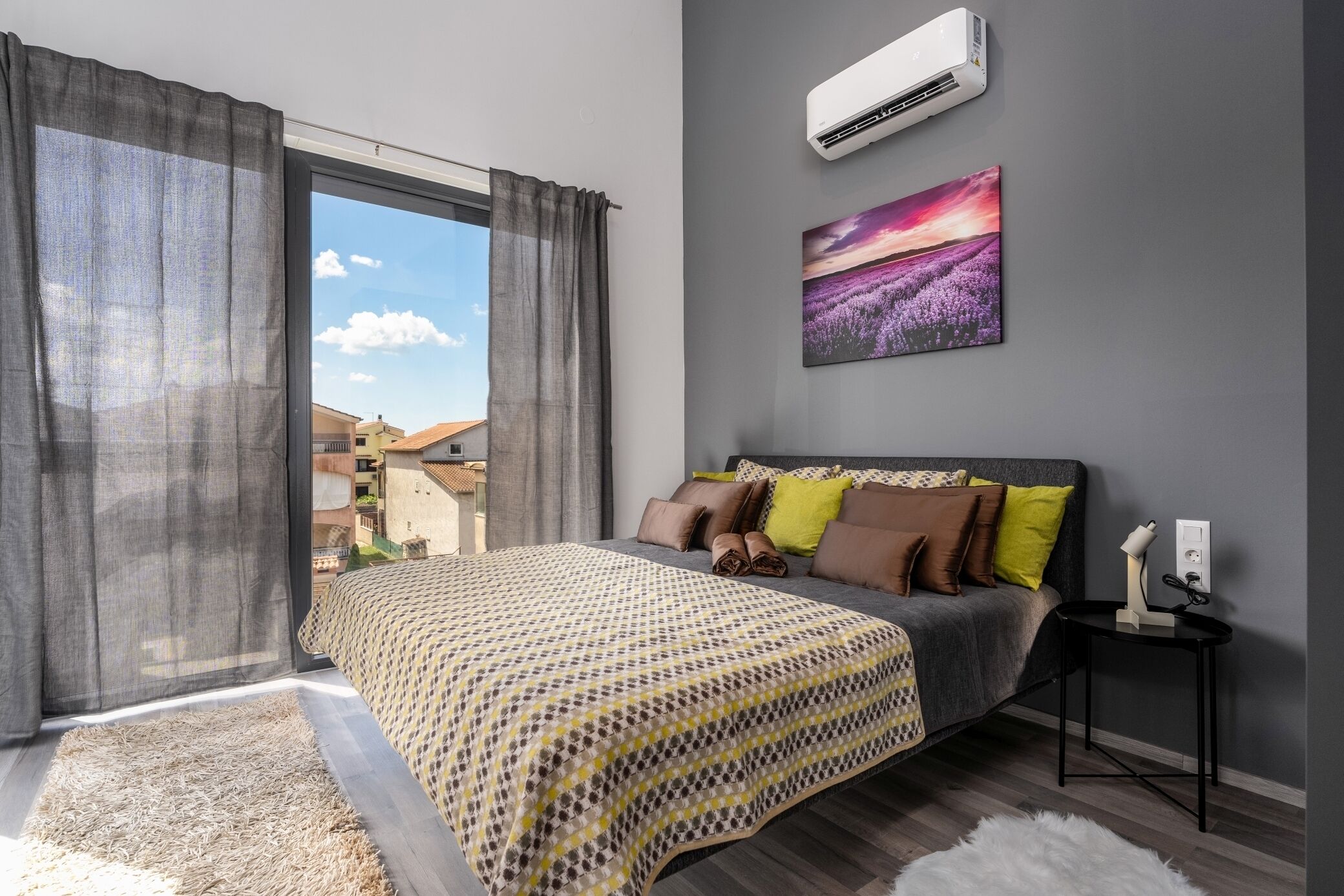 Camera da letto con un letto grande, tende grigie, vista sugli edifici, condizionatore d'aria e un quadro appeso al muro.