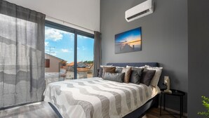 A modern bedroom with a large bed, a view of the sky through a window, and an air conditioning unit on the wall.