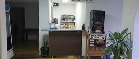 Ein Empfangsbereich mit Theke, einem Schild mit der Aufschrift 'RECEPCIÓN', einer Kaffeemaschine, einer Pflanze und einem Schild, das 'VENTA DE CAZUELAS DE FRUJOLES' bewirbt.