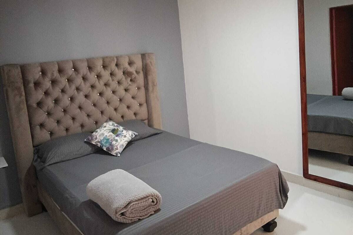 A bedroom with a tufted headboard, a grey bedspread, a rolled towel, and a mirror on the wall.