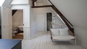 A room with a sloped ceiling, a white door, a white bench, and a visible bathroom with a toilet and bathtub.