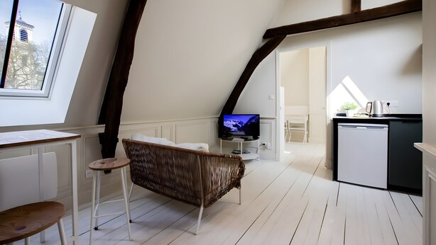 A loft-style room with a television, a wicker chair, a small table, and a kitchen area.