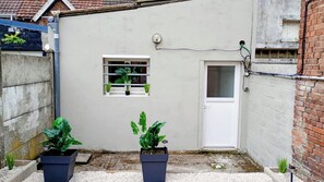 A small courtyard with a white door, a window with plants, and two potted plants.