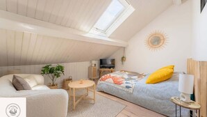 A cozy loft bedroom with a sloped ceiling, a skylight, a bed with a colorful blanket, a sofa, a small round table, and a TV. - Les combles d'Eugene, proche Bollaert, 2pers (Lens)
