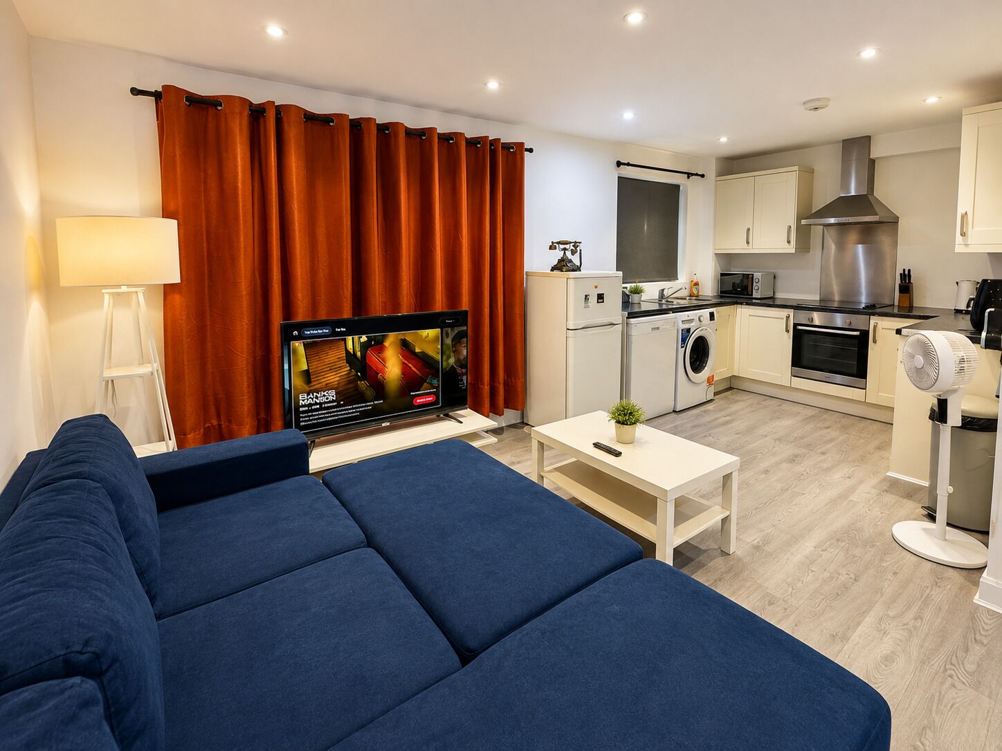 A modern living room with a sofa, a flat-screen TV, a coffee table, and a kitchen area with appliances.