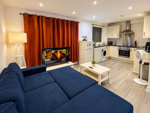 A modern living room with a sofa, a flat-screen TV, a coffee table, and a kitchen area with appliances. - Stylish 2-Bed Ground-Floor Flat | Wimbledon & Southfields | Sleeps 6 (London)