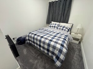 A bedroom with a bed, a nightstand, a lamp, a power strip, and a bag on the floor.