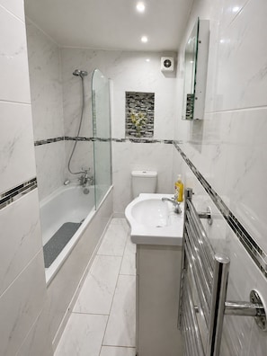 Une salle de bain moderne avec une douche à parois de verre, un lavabo blanc avec un miroir au-dessus et des toilettes dont le mur arrière est recouvert de mosaïques.
