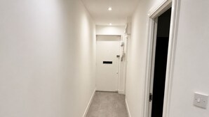A narrow hallway with a door at the end, a light switch on the wall, and a vent on the ceiling.