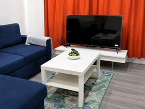 Un salon moderne avec un canapé bleu, une table basse blanche ornée d’une plante et un téléviseur à écran plat posé sur un meuble.
