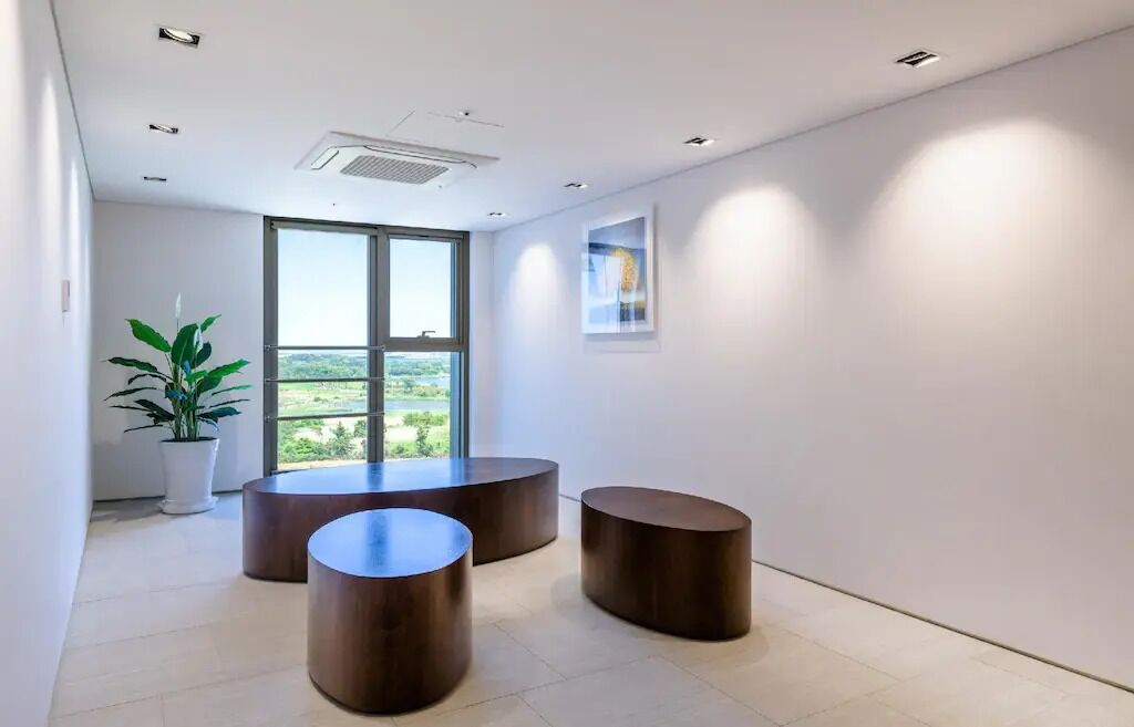 Un interior moderno con tres mesas redondas de madera, una planta en maceta y un ventanal con vistas a zonas verdes.