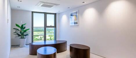 Un interior moderno con tres mesas redondas de madera, una planta en maceta y un ventanal con vistas a zonas verdes.